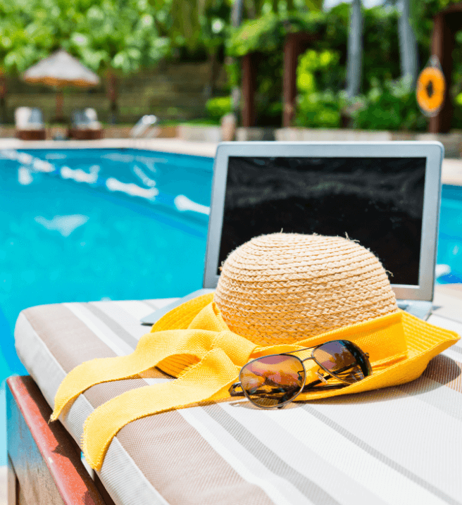 Photo of a straw sunhat, sunglasses, and a laptop sitting on a sunbed next to the pool.